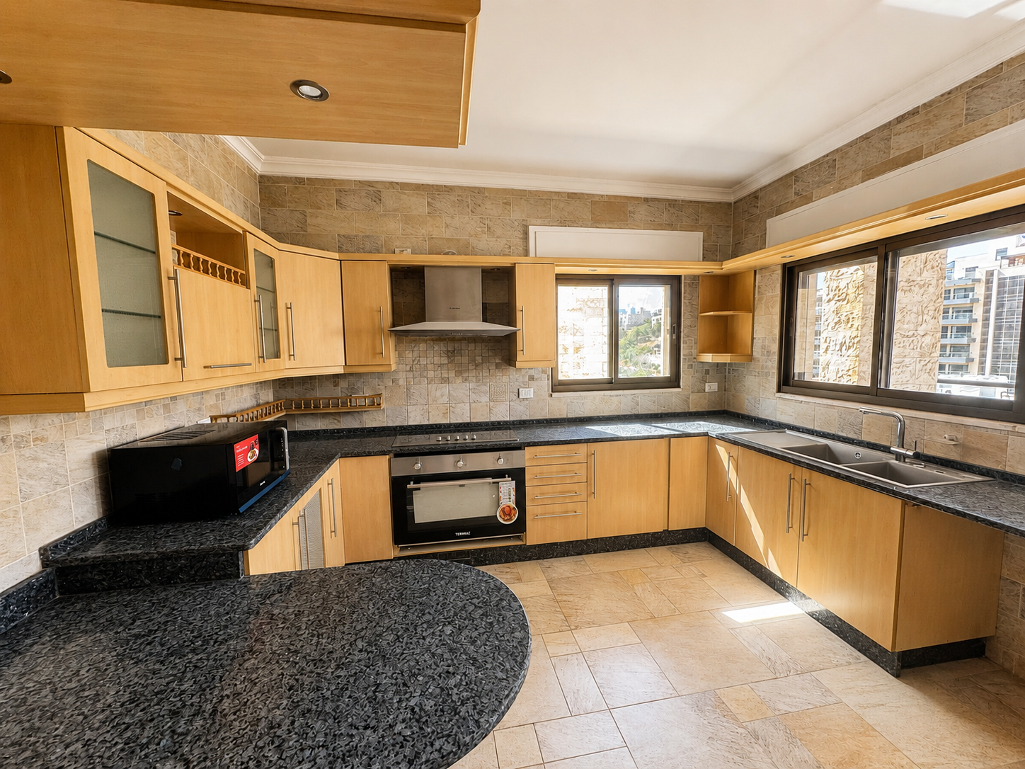 36e7d43c-7c1a-48ed-8f6f-784e661355a6 1 Modern L-shaped kitchen with light wood cabinets, black granite countertops, stainless steel oven and range hood, microwave, and a double sink under large windows.