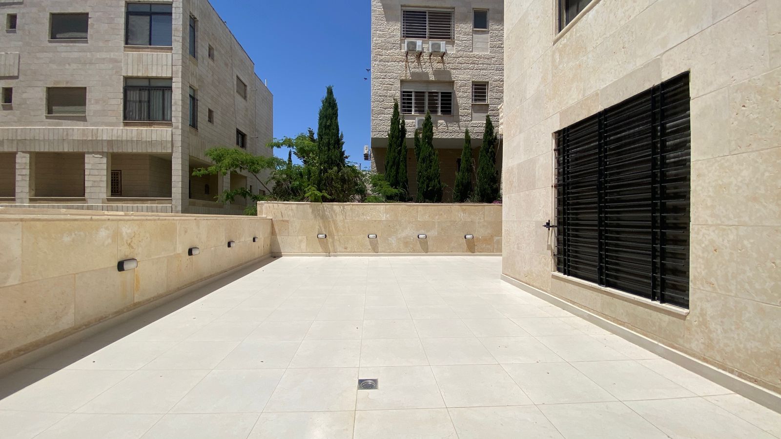 37541ae5-7b2f-4bec-a16e-fd00494643d6 Open concrete courtyard with beige stone walls, the right wall has a large barred window and door, center features a drain cap on tiled floor, blue sky above.