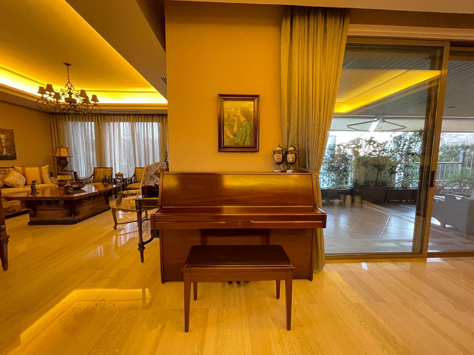 381179a3-aa6f-4f17-8e07-ccd5c9a8dc4c Upright wooden piano at center of a warmly lit living room, with a bench and a painting on the wall behind it, and a balcony door to the right.