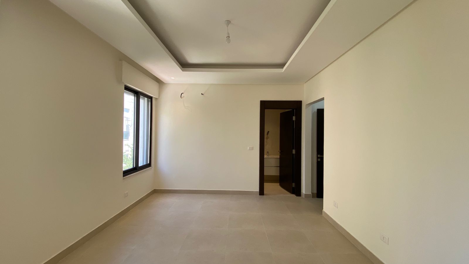 3a6e6a0c-ed7d-4566-803d-a3f7db6a8e26 Empty living area with beige walls, large left window, light tiled floor, and a shallow tray ceiling with a hanging bulb.