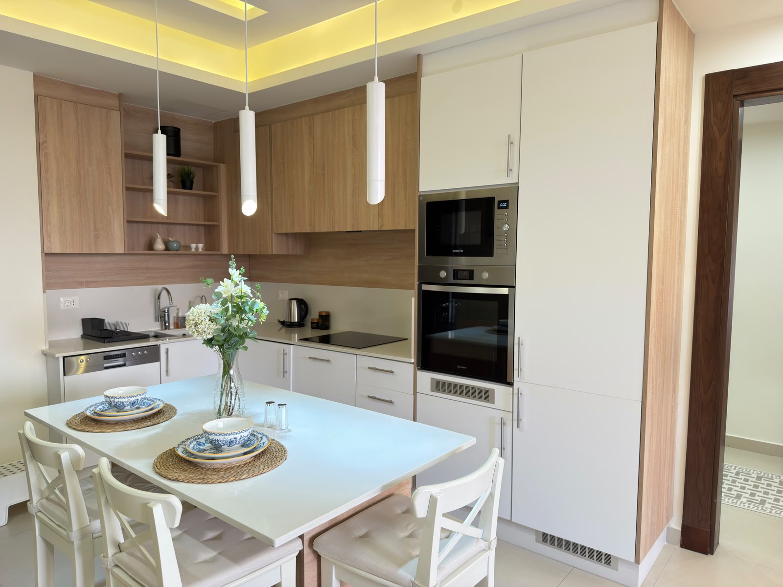 3b242947-bf97-4447-a2ec-6364e7ee0b85 Modern kitchen with white and wood cabinets, pendant lights, and a light-blue island table set for four with bowls and plates.