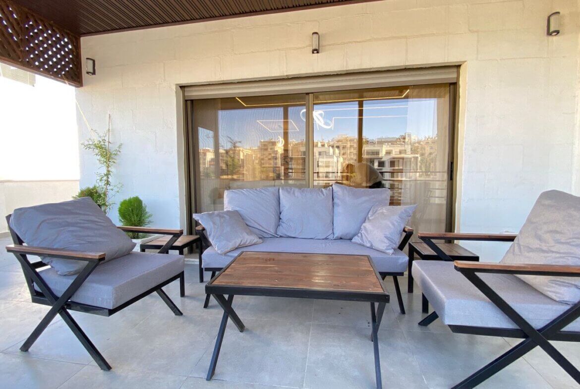Outdoor patio with gray cushioned sofa and matching armchairs around a wooden coffee table on a tiled deck beside sliding glass doors.