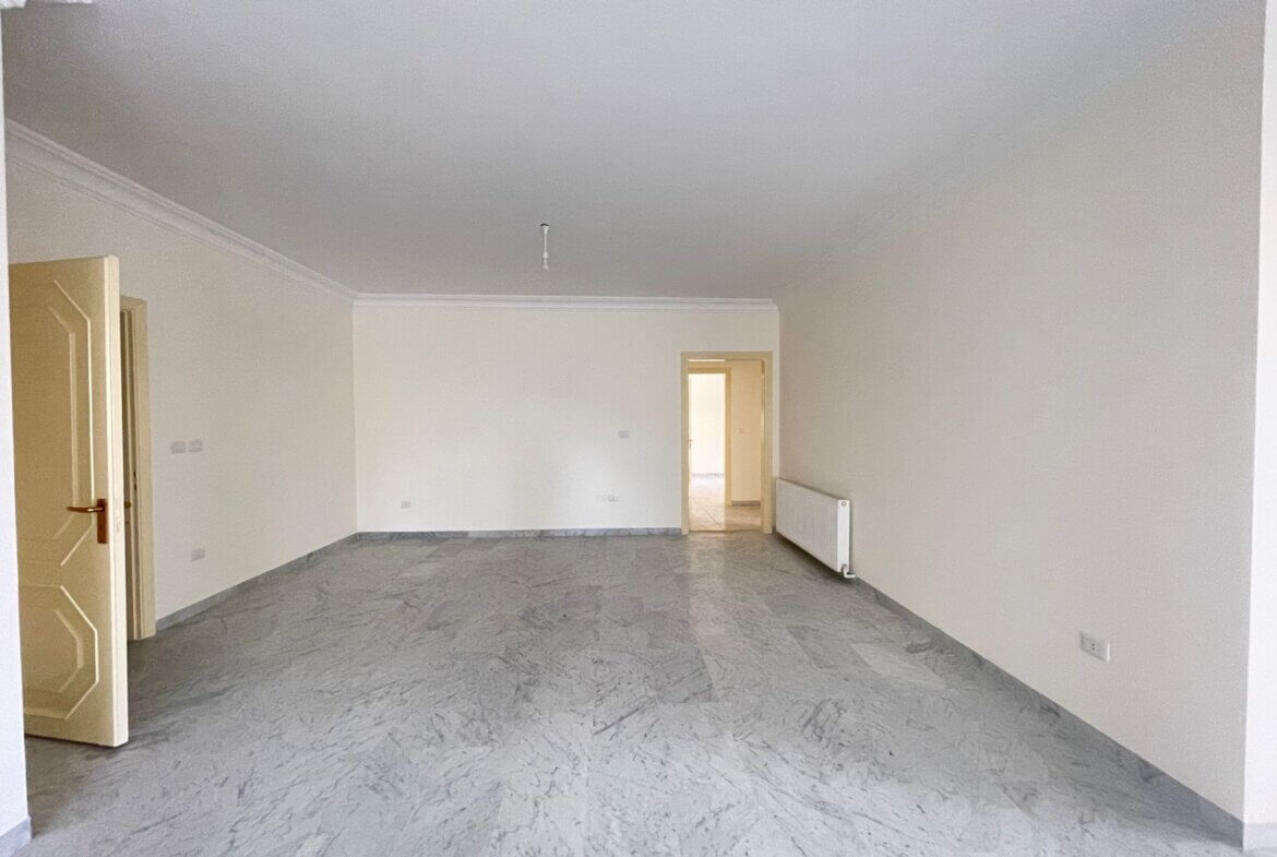 3dbcb7af-0beb-4a08-baae-1333513cca92 Empty rectangular living room with light-colored walls, marble floor, and an open door on the left; radiator on the right wall and another doorway in the back.