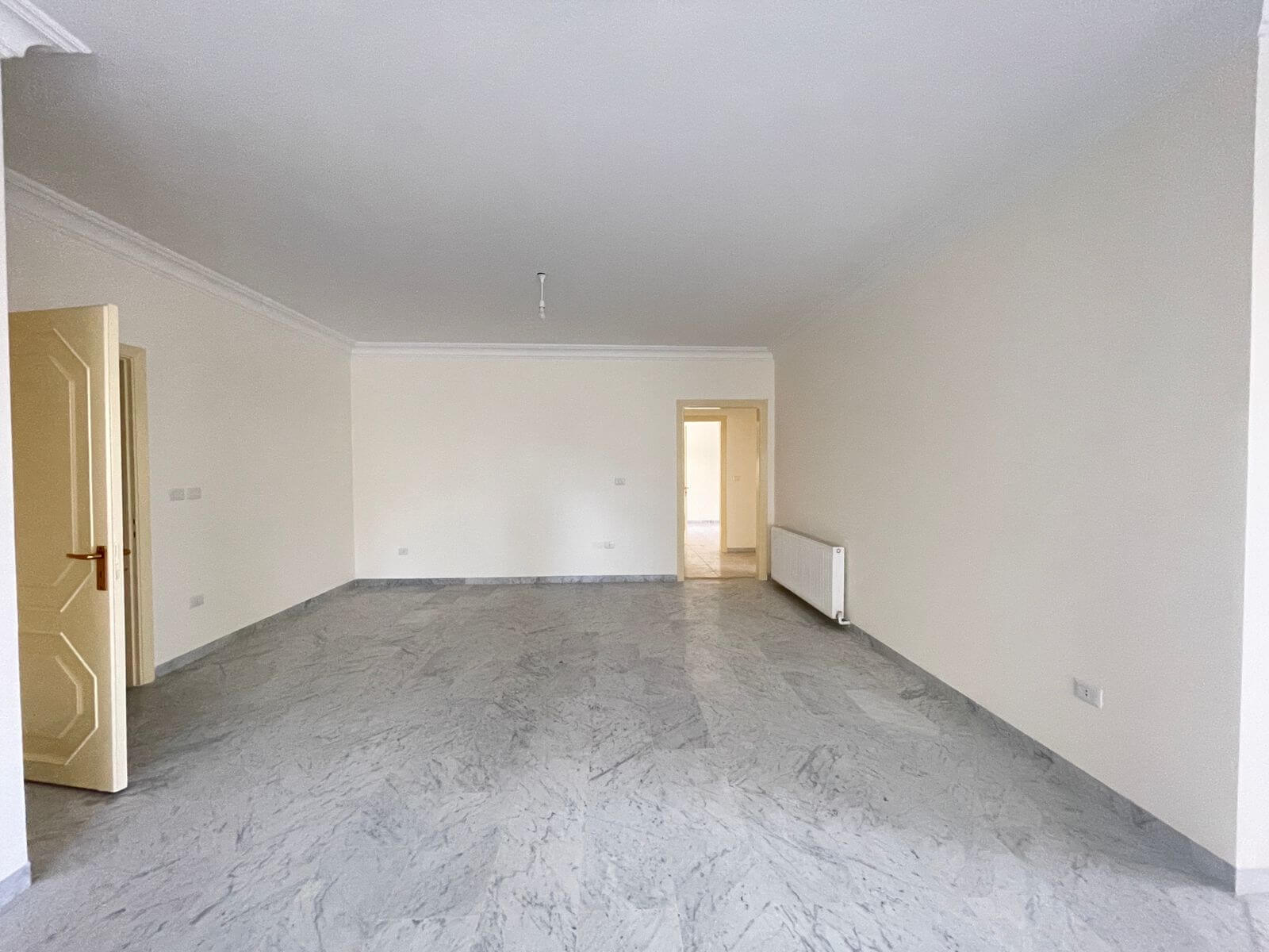 3dbcb7af-0beb-4a08-baae-1333513cca92 Empty rectangular living room with light-colored walls, marble floor, and an open door on the left; radiator on the right wall and another doorway in the back.