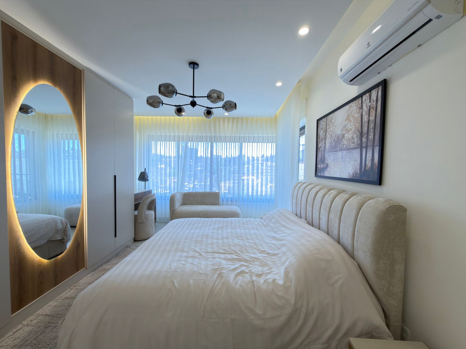 3e2ddcb1-f3bc-4cf1-b049-4a71b3a9abf1 Modern bedroom with white bedding and a beige tufted headboard, circular backlit mirror on a wood panel, and a white wardrobe.