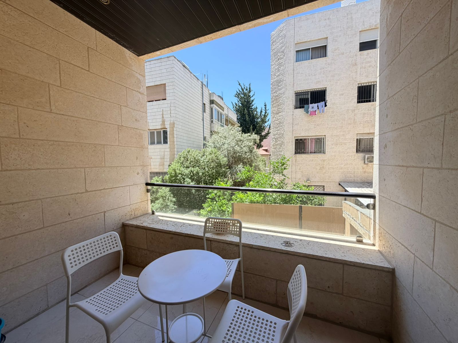 3e973fdf-8188-46de-aaa7-300ff6b47847 Balcony seating area with a round white table and four perforated chairs, overlooking beige apartment buildings and trees.