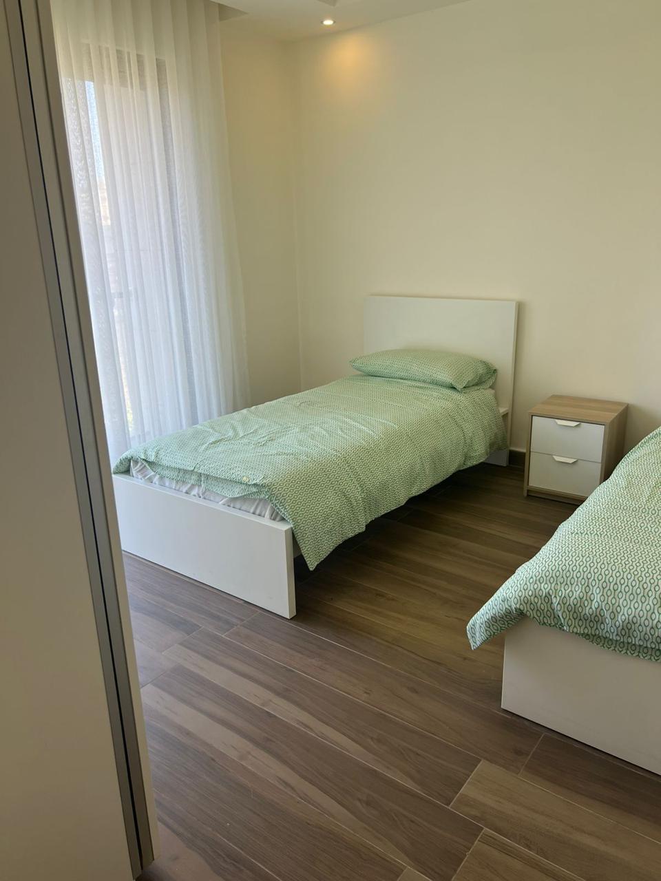 4138a1d4-2ef4-4bd7-b3ac-a79f6b7a28c4 Small bedroom with two single beds in mint bedding, white headboards, and a wooden nightstand between them or nearby.