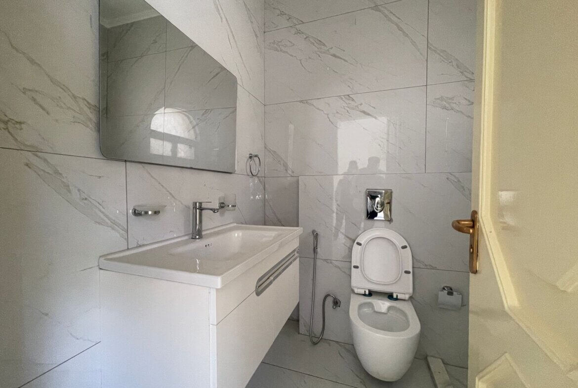 4196cb05-d8fd-49c1-93d0-3cd6cf49559c Modern bathroom with white marble tiles, wall-mounted vanity sink, large mirror, and a toilet with bidet hose nearby.