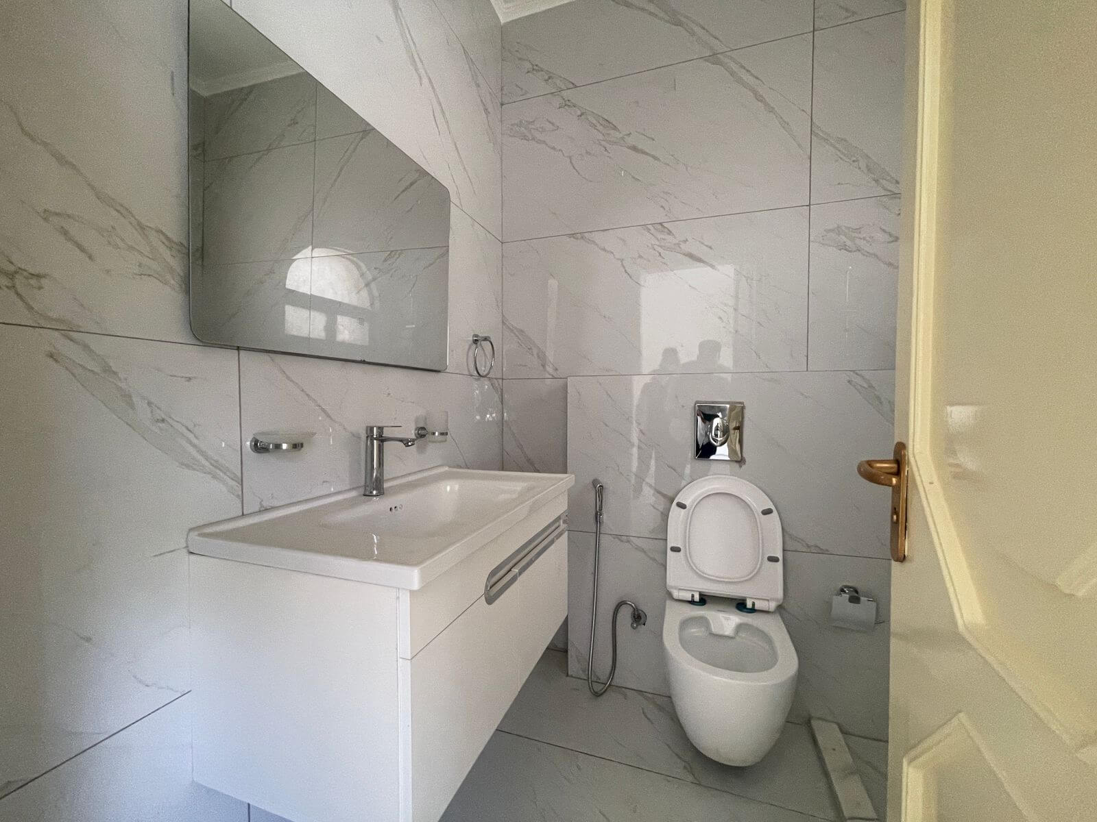 4196cb05-d8fd-49c1-93d0-3cd6cf49559c Modern bathroom with white marble tiles, wall-mounted vanity sink, large mirror, and a toilet with bidet hose nearby.