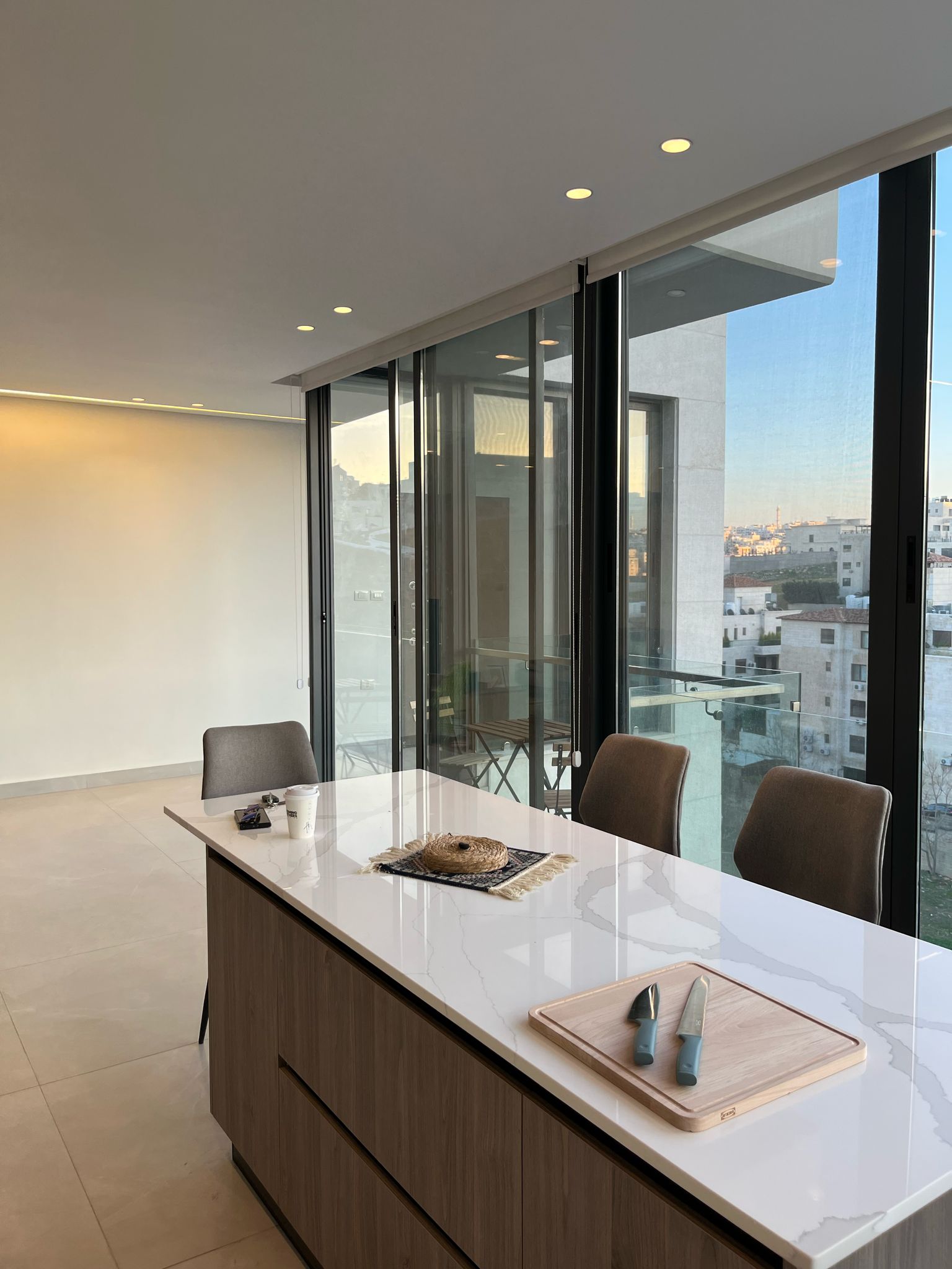 41bf46a2-3dc7-481b-8355-cbc06de31da3 Modern kitchen island with a white marble countertop, knives on a cutting board, and large glass windows overlooking the cityscape across from beige chairs.