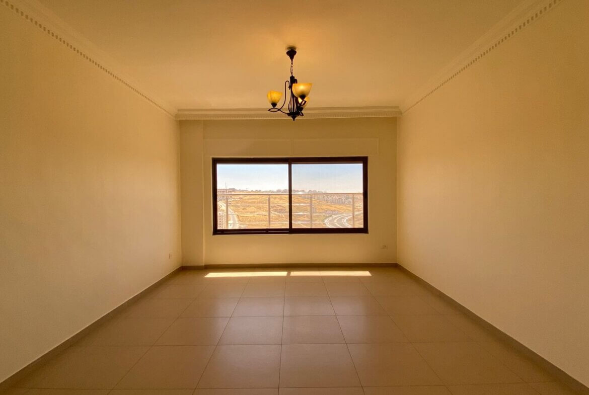 4207691f-5076-4bb5-b4ee-60553313eb1b Empty rectangular room with beige walls, tiled floor, and a chandelier; a large window reveals a dry, distant landscape outside the city.