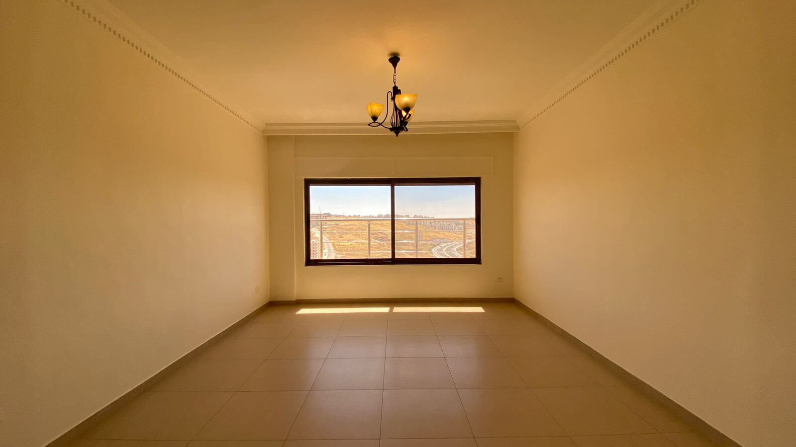 4207691f-5076-4bb5-b4ee-60553313eb1b Empty rectangular room with beige walls, tiled floor, and a chandelier; a large window reveals a dry, distant landscape outside the city.