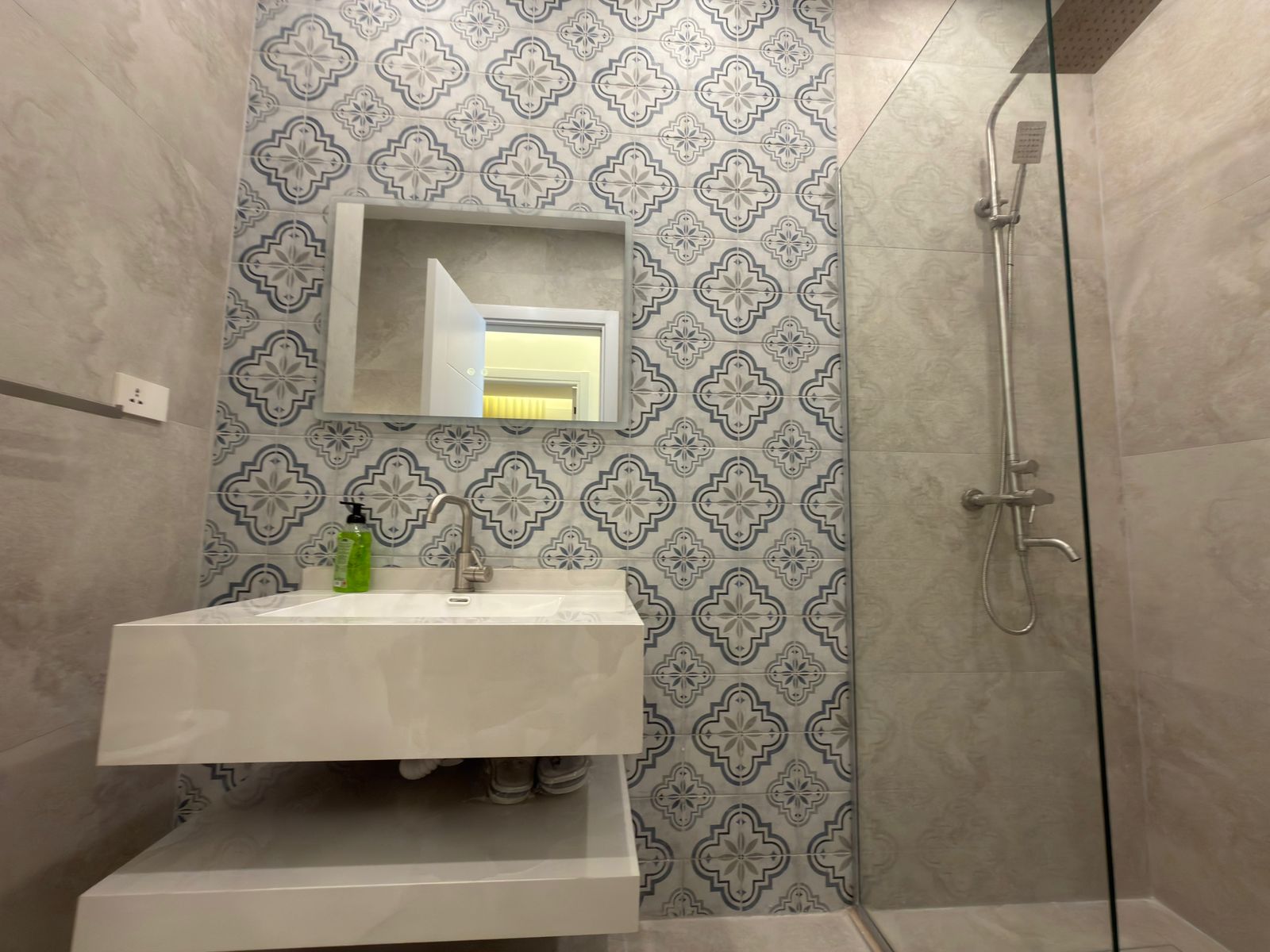43a53166-e14d-4c5f-a7b1-194beb776f55 Modern bathroom with a white floating vanity, green soap bottle, and a rectangular mirror above a patterned tile wall featuring blue-gray shapes.