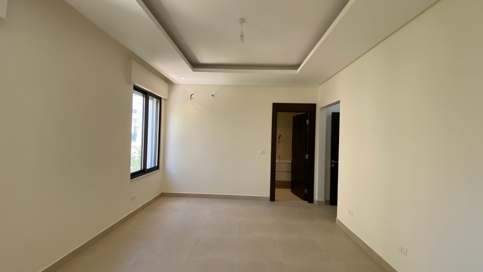 441600b5-765c-435e-9863-75ac81dd447a Empty living room with beige walls, a large left-side window, and a recessed tray ceiling; doorway to a bathroom on the far wall.