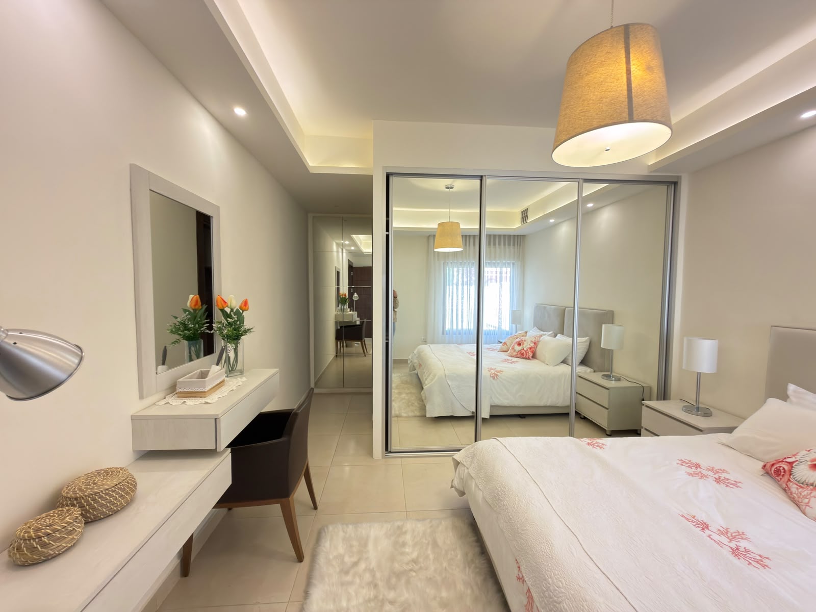 445291cd-c4d5-4e3a-8285-5ffdc44ebd33 Bright modern hotel bedroom with a white desk, mirror, and vase of orange-tipped flowers; mirrored closet doors reflect the bed and lampshade.