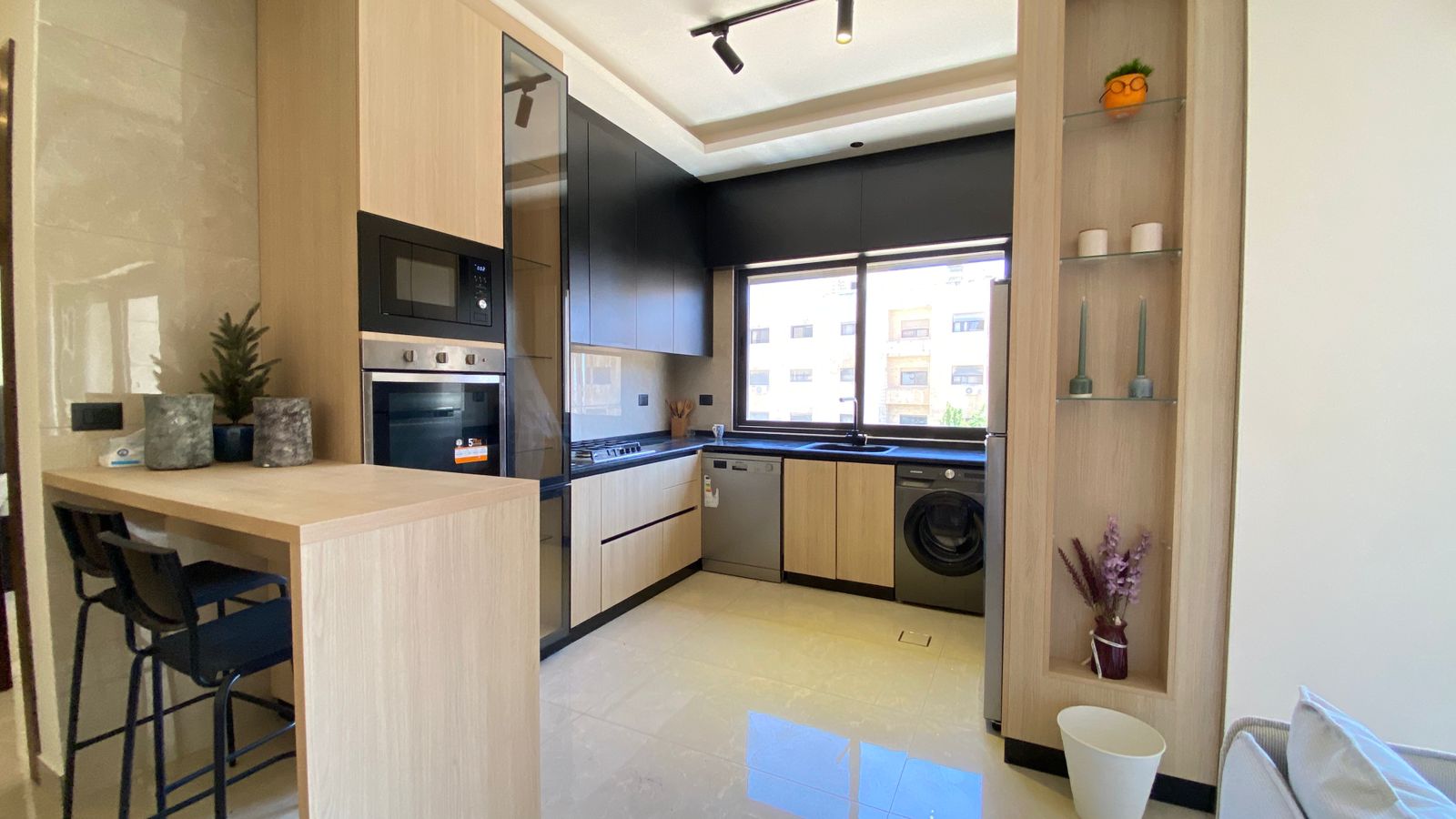 44a50aec-29bf-4259-881c-46b5fbdf575e Modern L-shaped kitchen with black upper cabinets, light wood lower cabinets, and stainless appliances under a large window overlooking buildings; island with two bar stools on the left