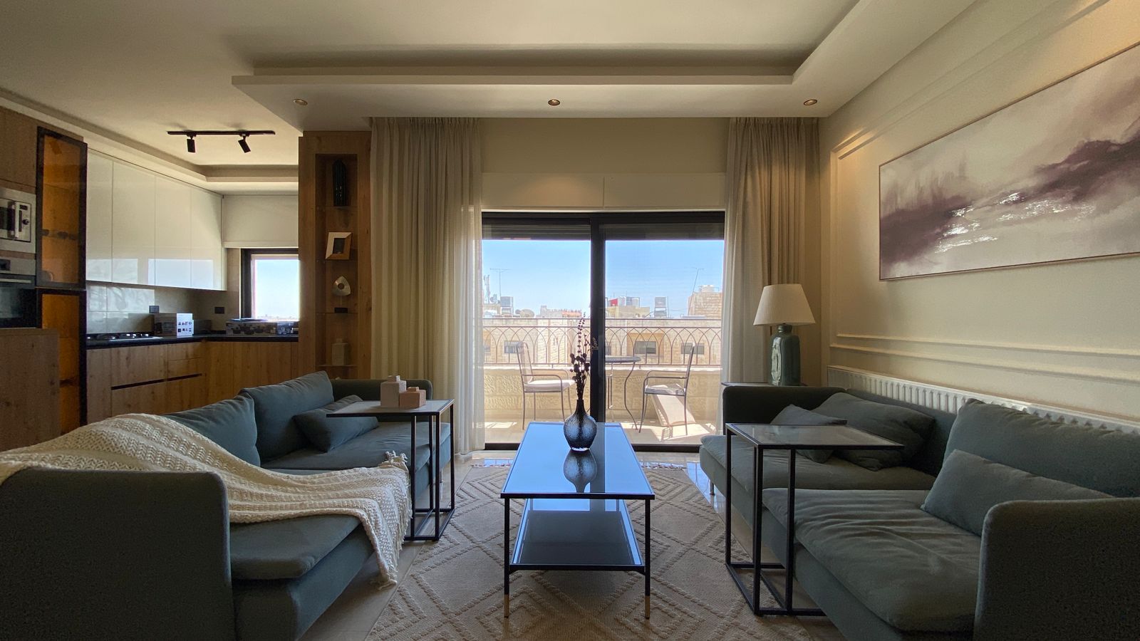 451fc88e-2278-4526-8b5c-00af6819a83d Living room with two blue couches facing each other, glass coffee table, and a balcony with city view through sliding doors on a bright day.