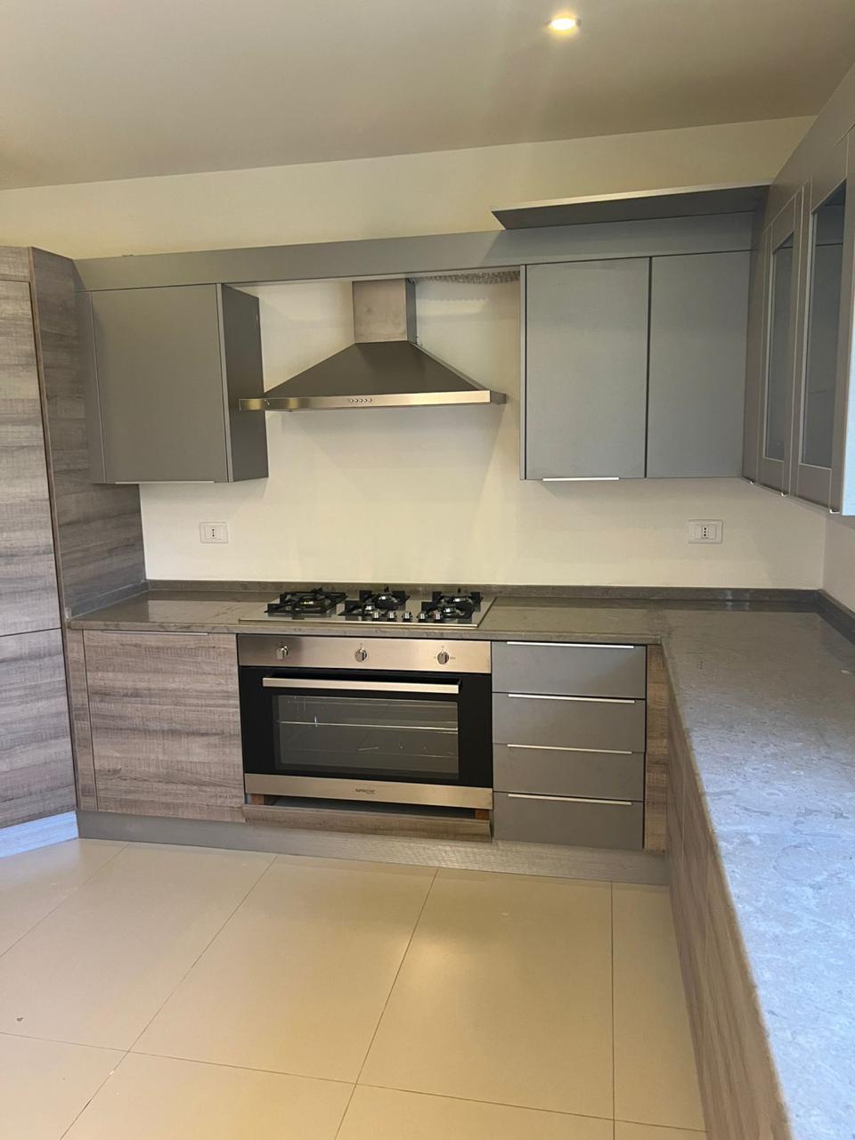 462ae9ed-0113-437a-9aec-0f740ec73109 Modern L-shaped kitchen with grey cabinets, stainless steel range and hood, and marble-like countertops.