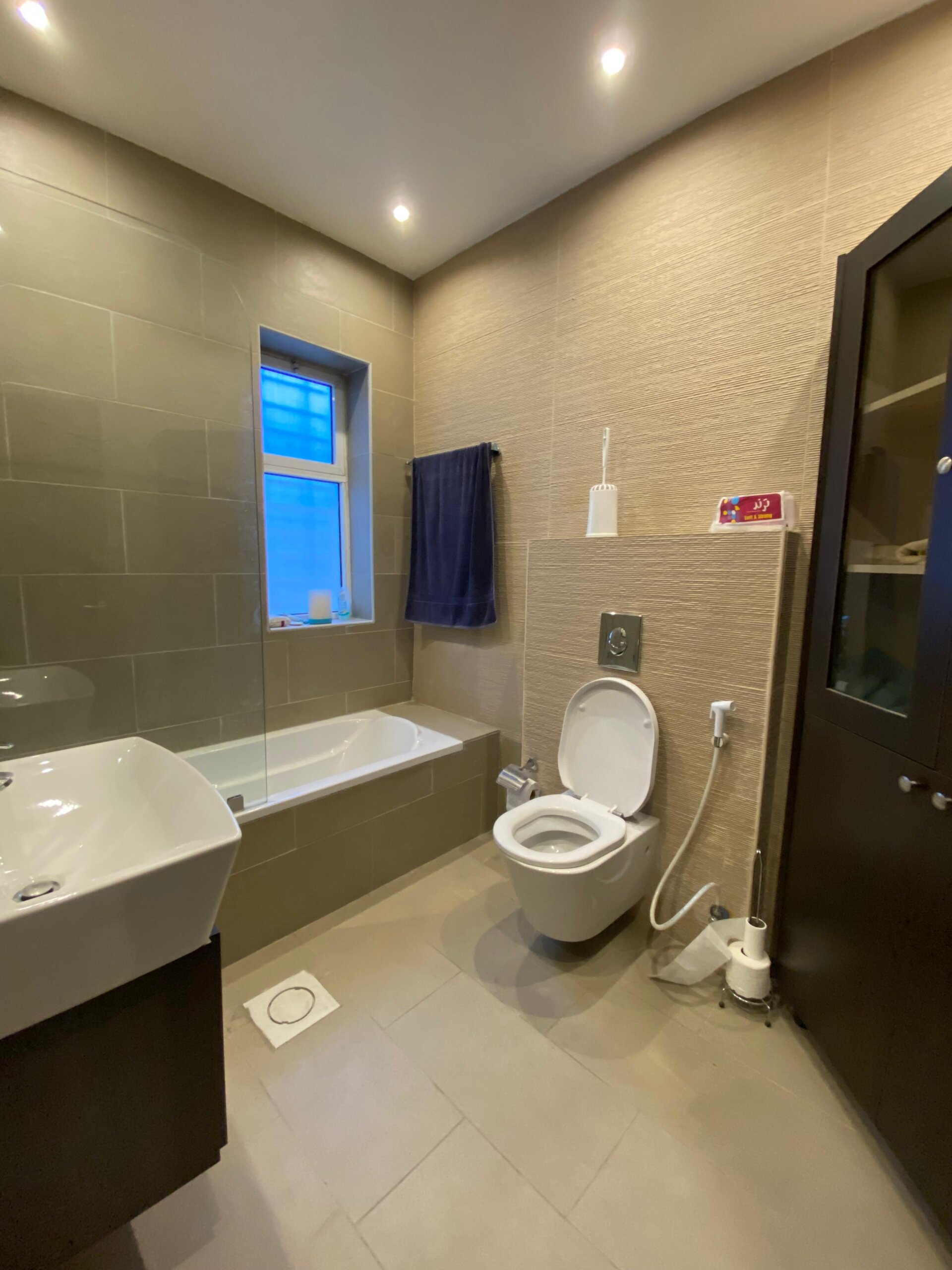 46e19dcf-98ac-42f7-9245-7246184af4f0 Modern bathroom with a bathtub along the left, sink, and beige tiled walls; a wall-mounted toilet with bidet spray nearby.