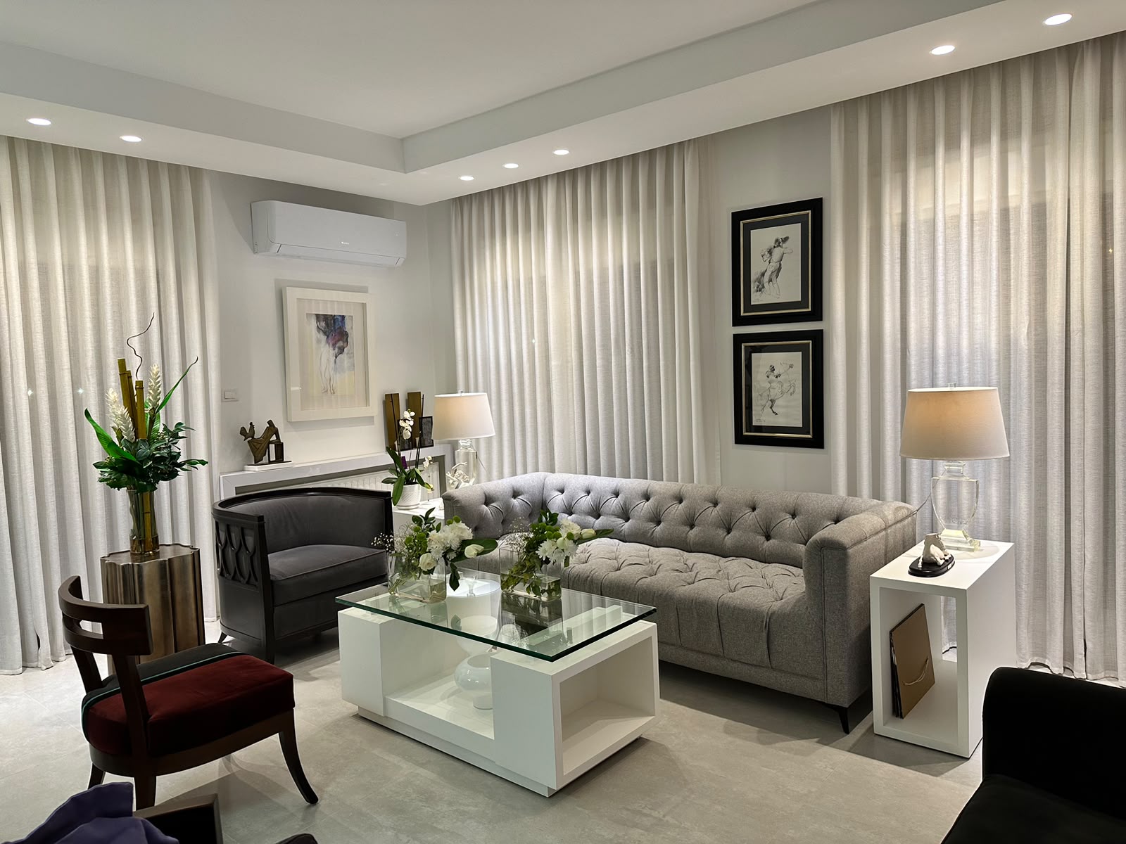4801c1b5-7dcb-4a85-b95a-19fffe05be7d Bright modern living room with a tufted gray sofa, glass coffee table, sheer white curtains, and soft ambient lighting.
