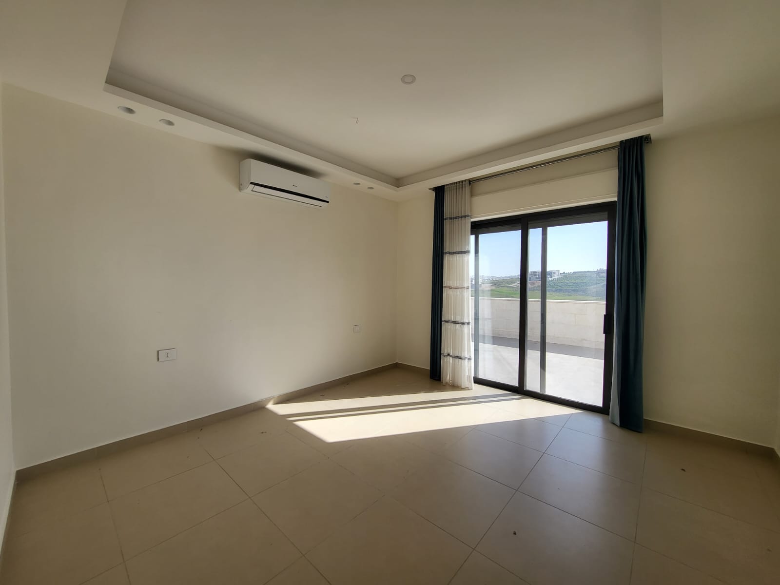 4a02cd61-a93d-4fb8-8815-470cc6f1e839 Empty living room with a large sliding glass door to a balcony, sheer and blue curtains, and a wall-mounted air conditioner.