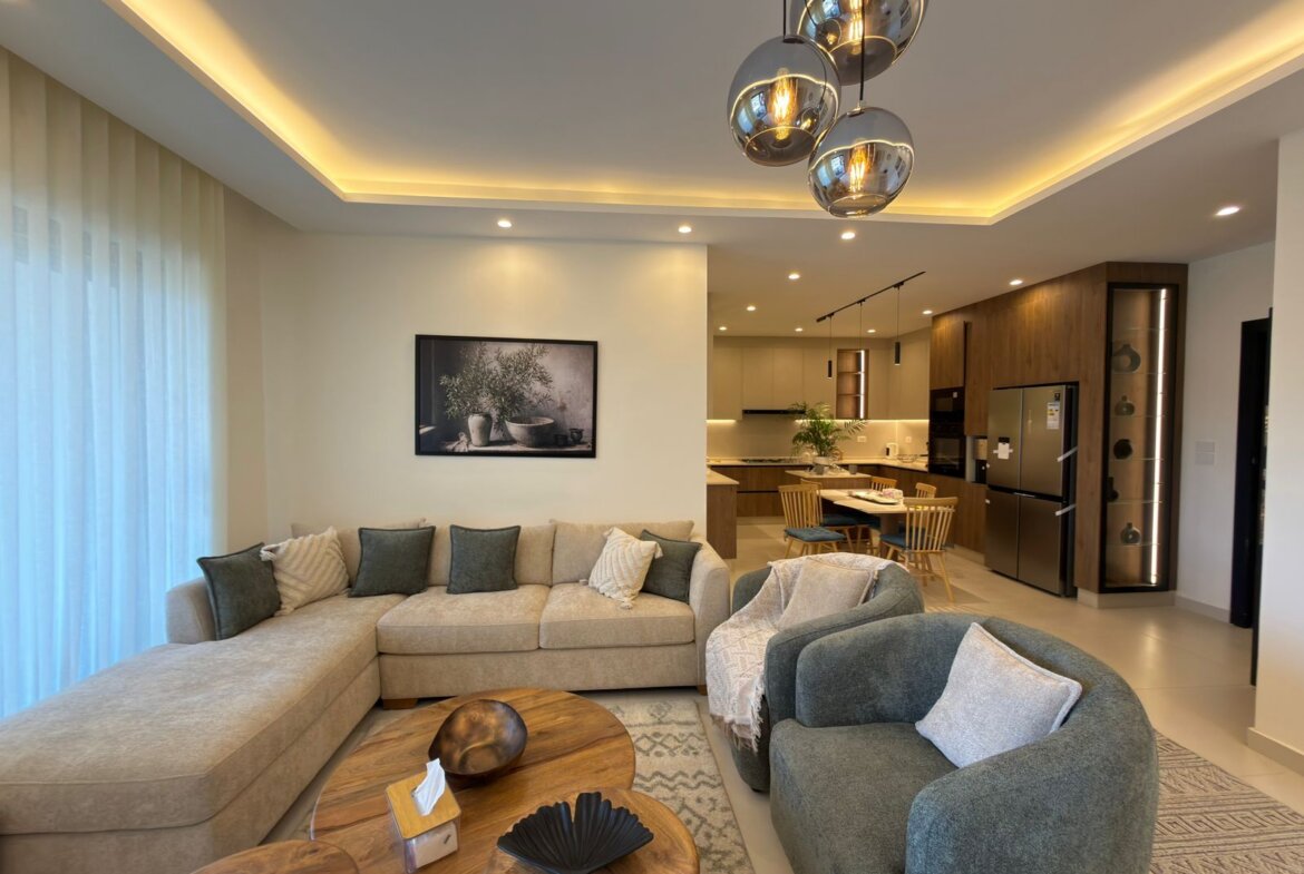 Open-concept living room with a beige sectional and gray cushions, nearby round wooden coffee tables, and a view into the modern kitchen.