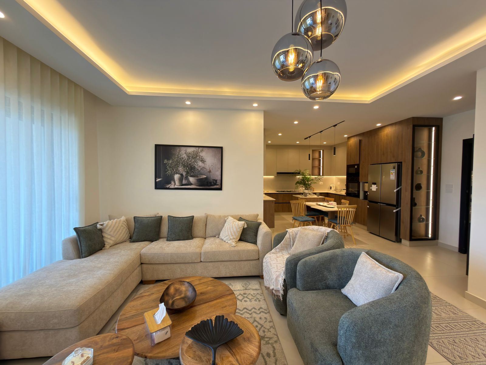 4d50f1a9-f662-4083-9a9e-b1543e9b544a Open-concept living room with a beige sectional and gray cushions, nearby round wooden coffee tables, and a view into the modern kitchen.