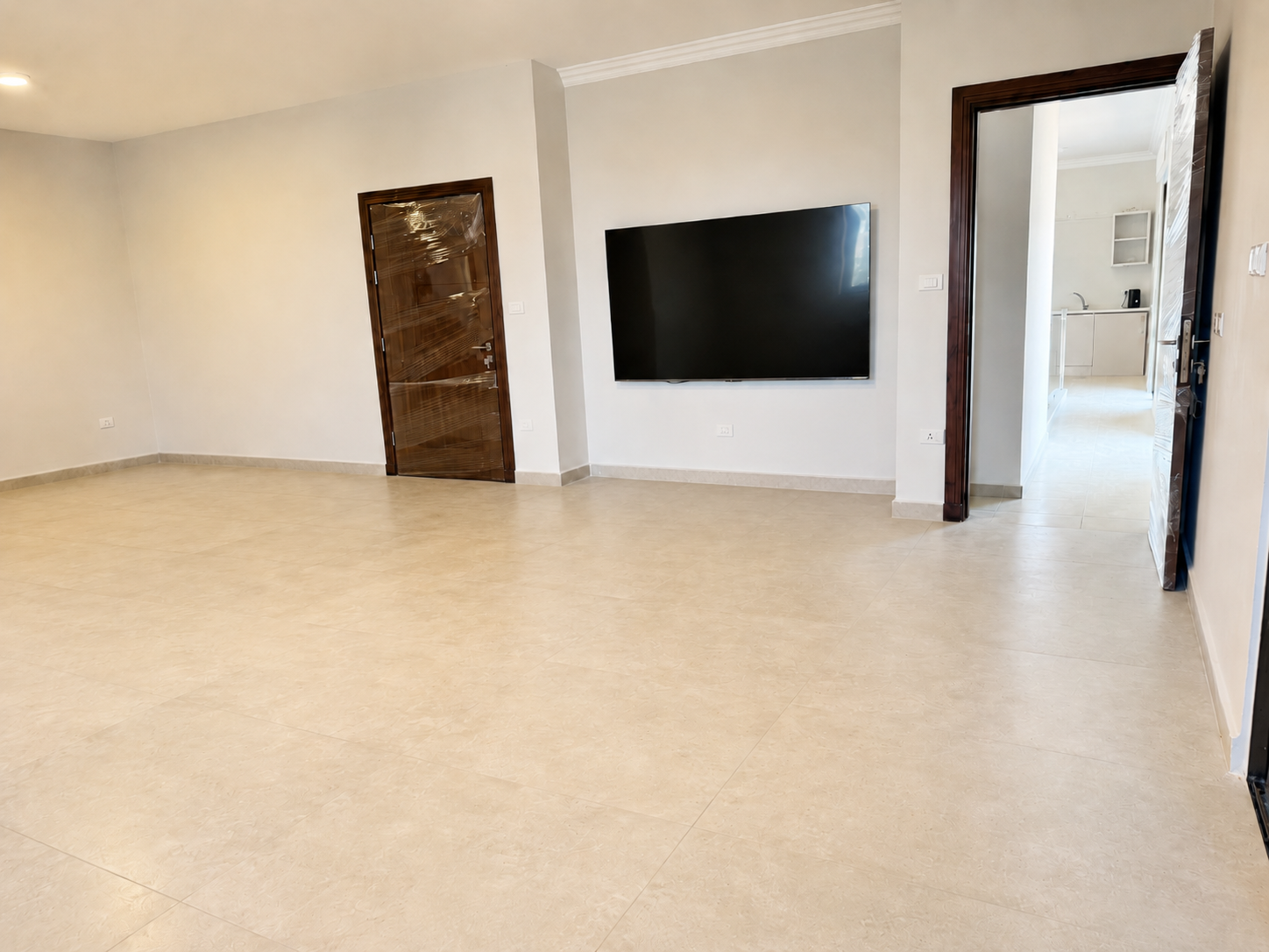 4e4a25e9-bdac-4164-8456-0171debf4253 Spacious living room with a wall-mounted flat-screen TV, beige tiled floor, and two wooden doors (left and right).