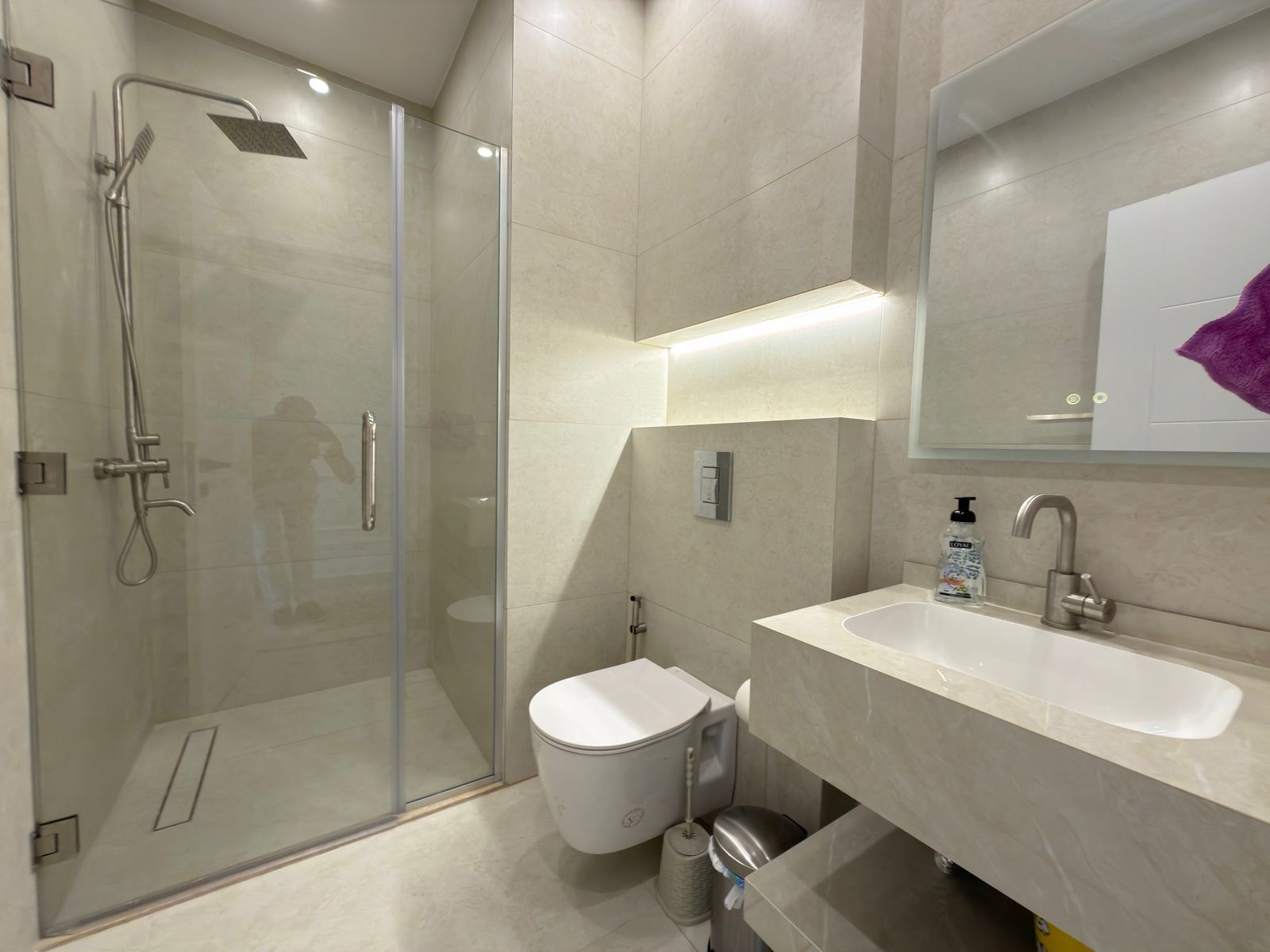 4e535968-769d-4607-9166-c75cb51de8e8 Modern beige-tiled bathroom with glass shower enclosure, wall-mounted toilet, and a white sink with brushed metal faucet.