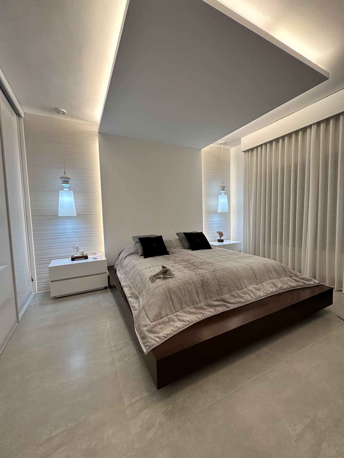 4f892184-ade8-4b76-a59f-5b64b9c17c28 Modern bedroom with a low dark wood platform bed, beige quilted duvet, two white nightstands, and pendant lights casting a soft glow; floor-to-ceiling curtains in neutral tones.