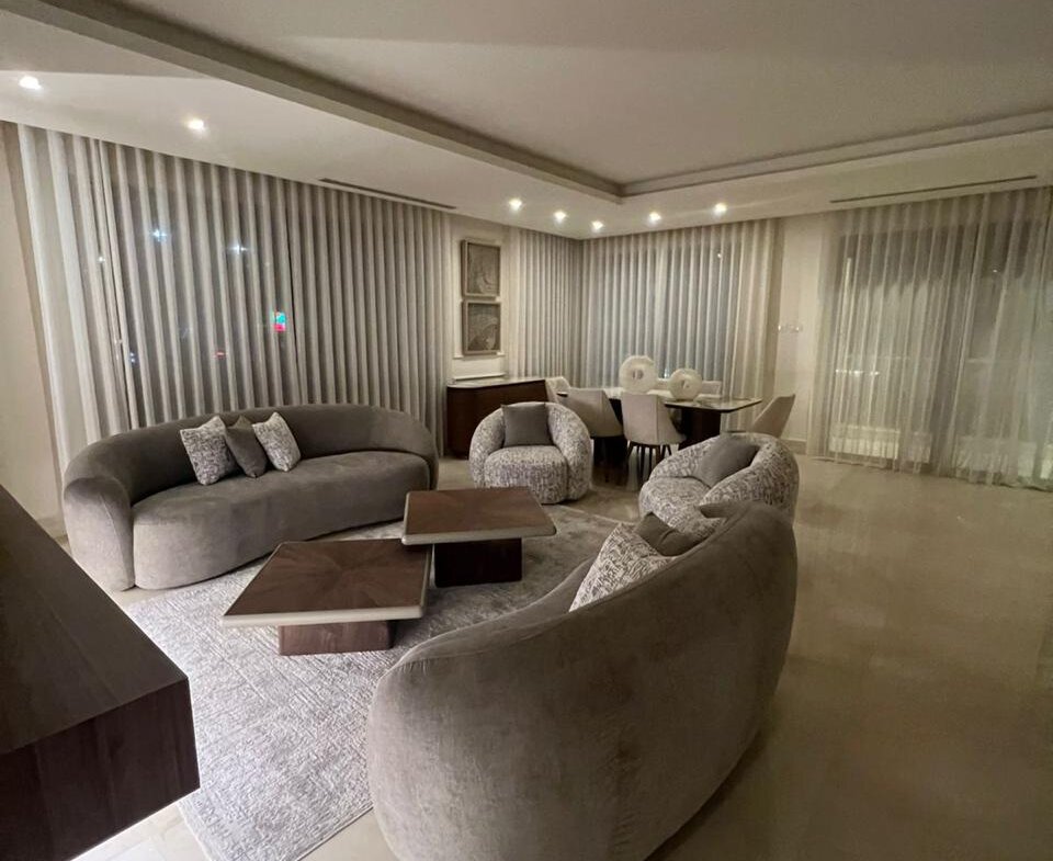 Modern living room with curved gray sofas around two wooden coffee tables, a light rug, and sheer-curtained windows in the background.