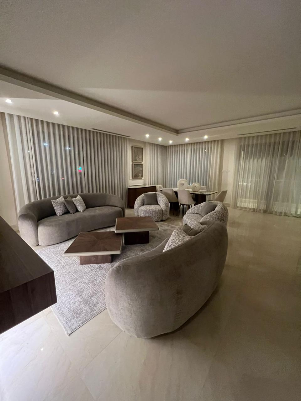 502ec918-3792-4cf0-90ad-67ecab2c9376 Modern living room with curved gray sofas around two wooden coffee tables, a light rug, and sheer-curtained windows in the background.