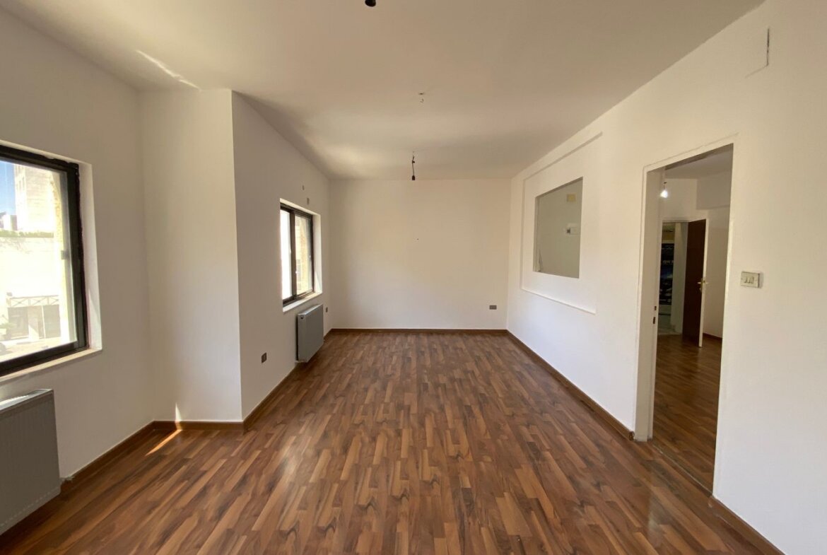 Empty rectangular room with wood-look flooring, white walls, and two large windows letting in sunlight