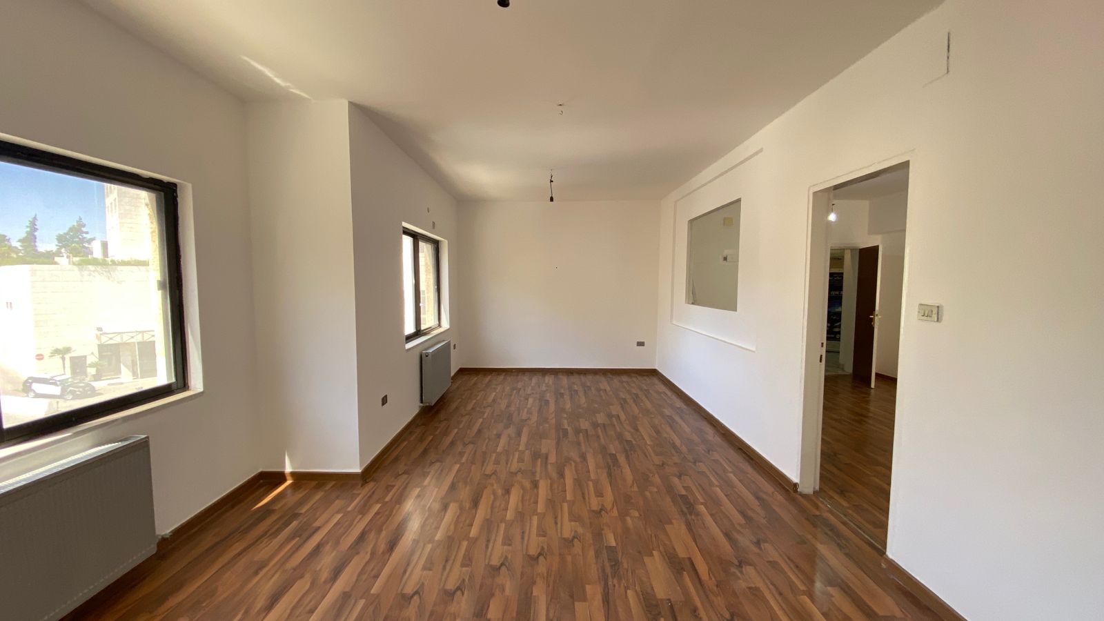 5072ef57-633d-4c32-9661-ead46028711f Empty rectangular room with wood-look flooring, white walls, and two large windows letting in sunlight