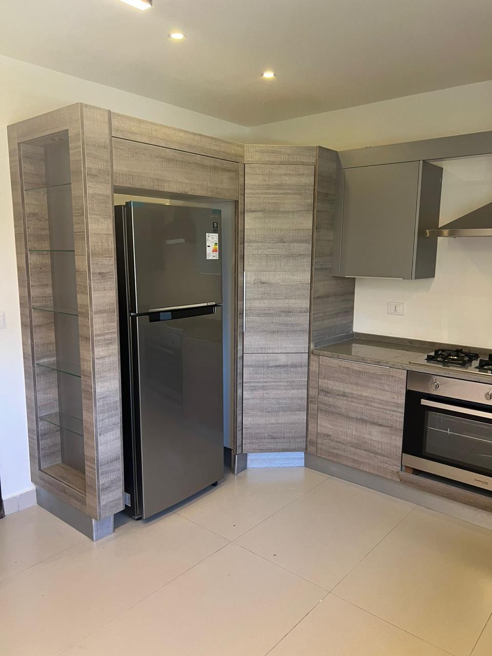 50b5747c-f62a-4bfd-bef2-57d8ef15dc07 Modern L-shaped kitchen with gray wood grain cabinets, stainless steel fridge, and built-in oven and gas cooktop bane to the right
