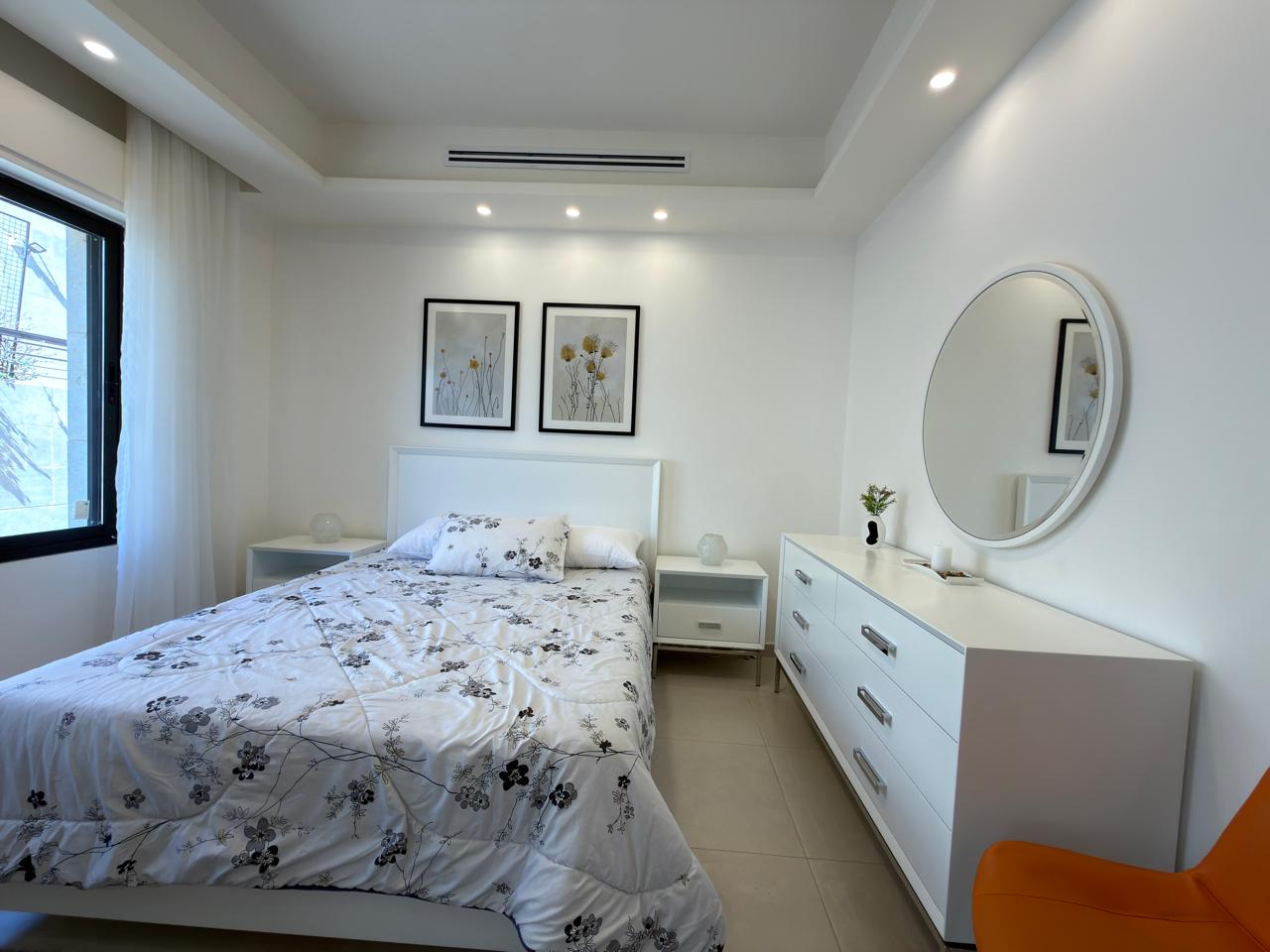 50d70de3-cc24-4317-a6f1-21a6f5a717aa Bright white bedroom with a floral quilted bed, two white nightstands, and a white dresser with a round wall mirror.