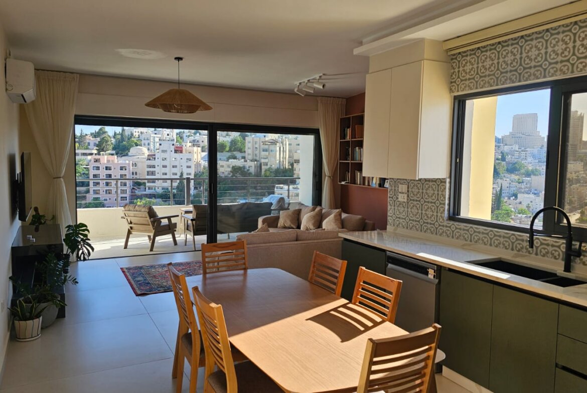 Open-plan living area with a wooden dining table, beige sofa, and kitchen along the right, opening to a balcony with city views.