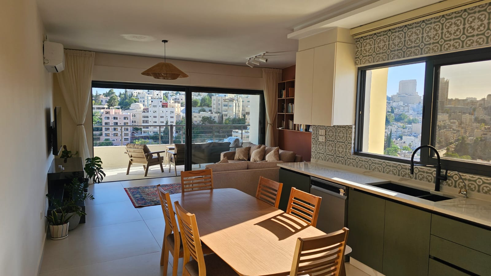 52925f1f-5b20-4d41-865b-49afa07879b2 Open-plan living area with a wooden dining table, beige sofa, and kitchen along the right, opening to a balcony with city views.