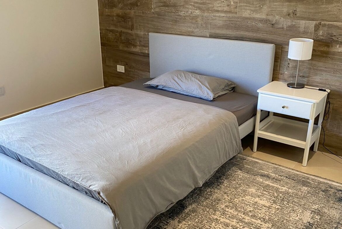 Bedroom with gray upholstered bed against a wood-paneled accent wall; white nightstand and lamp to the right.
