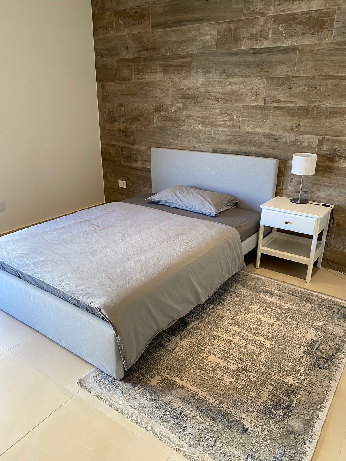 529ff6c3-f87c-424e-ad90-2492b1f29d8b Bedroom with gray upholstered bed against a wood-paneled accent wall; white nightstand and lamp to the right.