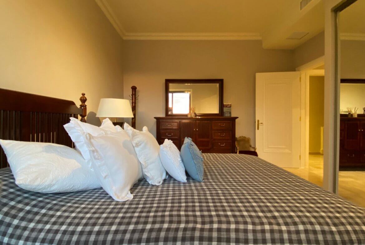 53a9489e-94ad-4156-913b-b45106e0b878 Bedroom with a large wooden bed, white pillows, and a blue-gray checkered duvet; dresser, mirror, and lamp in the background.