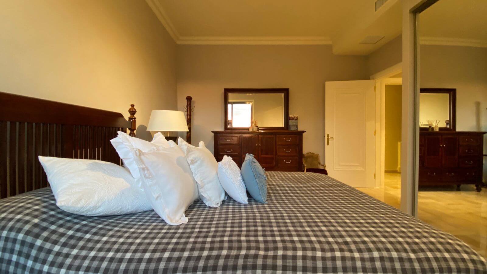 53a9489e-94ad-4156-913b-b45106e0b878 Bedroom with a large wooden bed, white pillows, and a blue-gray checkered duvet; dresser, mirror, and lamp in the background.