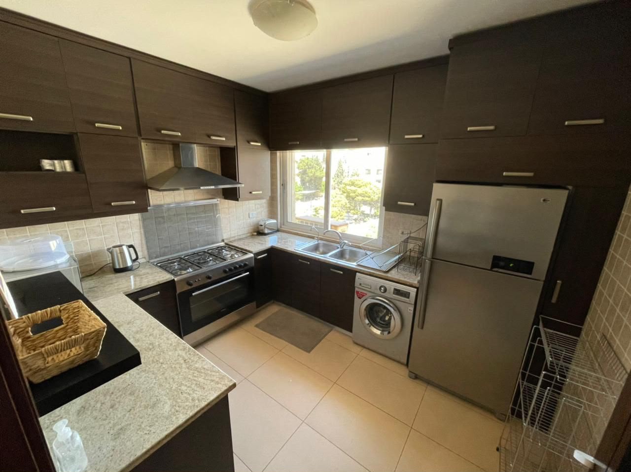 5454c5e2-ec37-4bf5-9823-98f3ff619fc7 U-shaped kitchen with dark wood cabinets, stainless steel fridge and washing machine, gas range with hood, double sink by a window, dish rack, and beige tile backsplash; basket on counter.