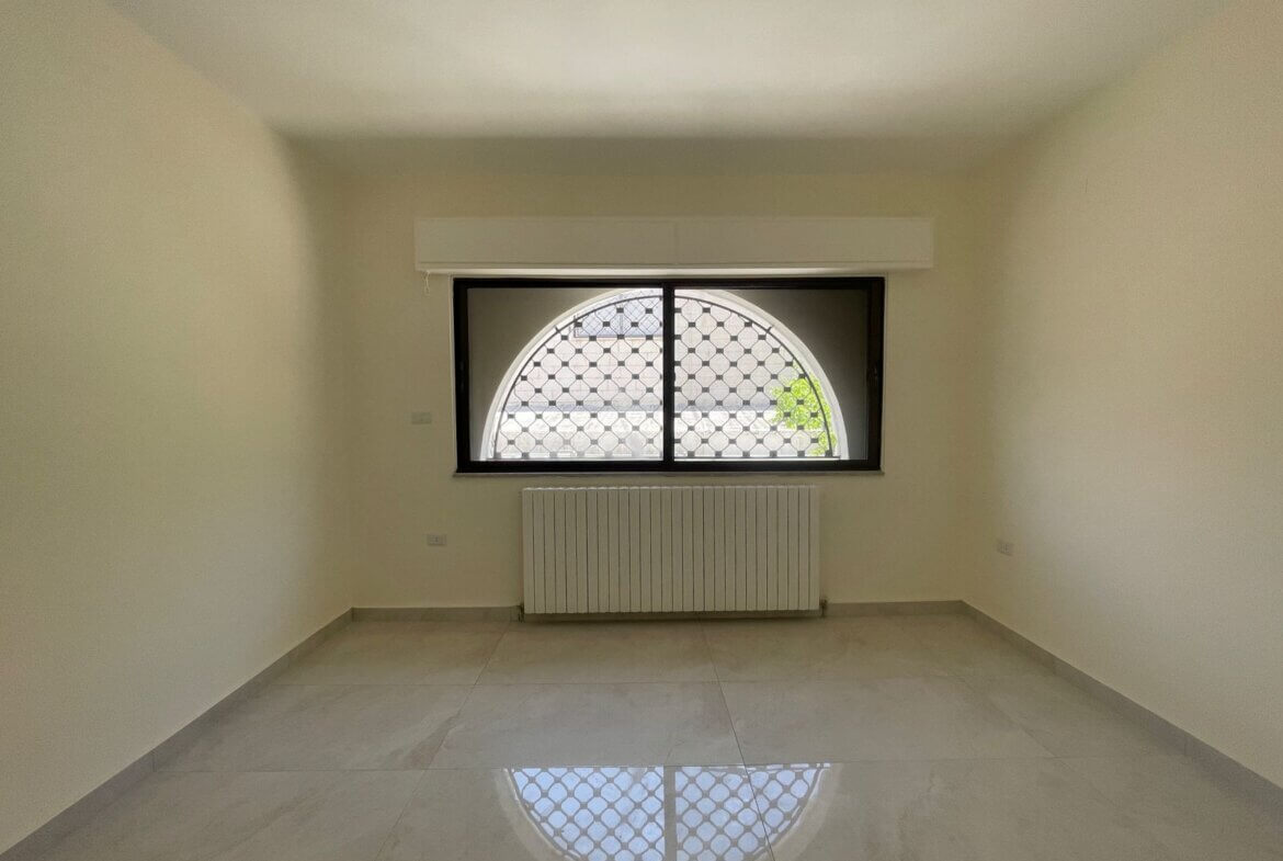 54c0b891-bb22-469c-b4d6-ad9fc9328798 Empty rectangular room with pale walls, arched window with a metal grid and a white radiator beneath, reflections on a glossy tiled floor.
