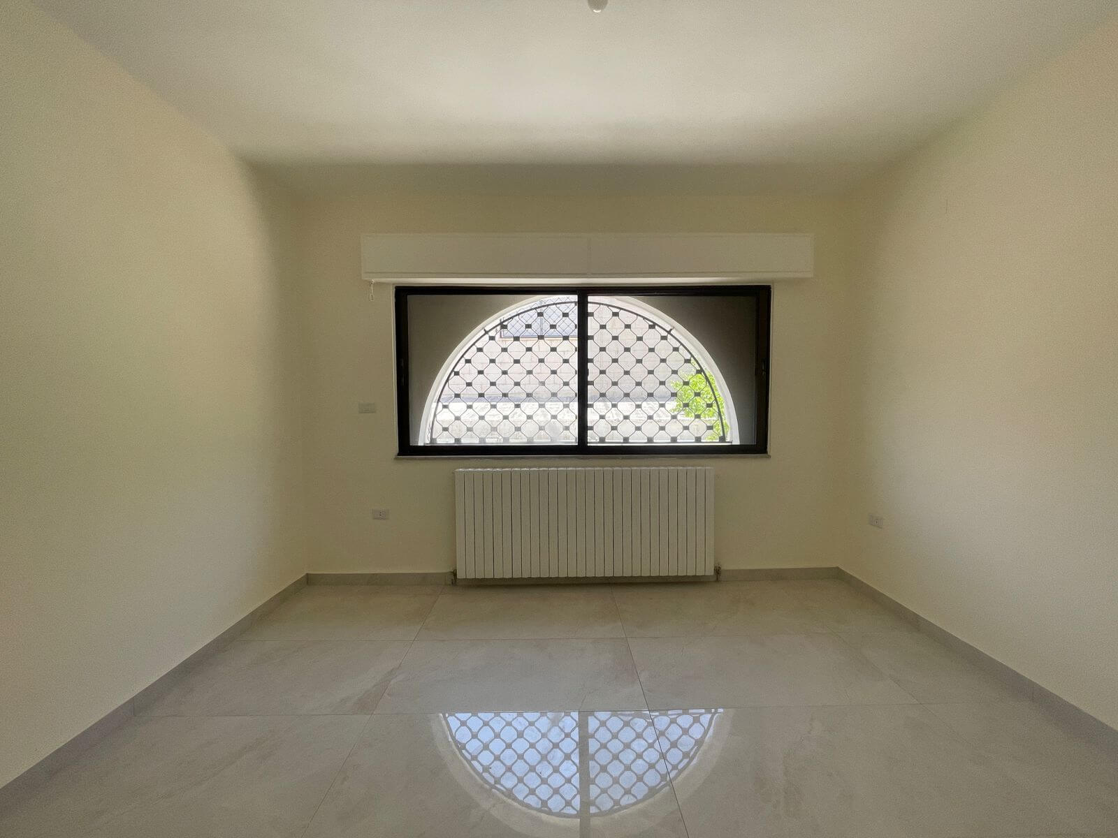 54c0b891-bb22-469c-b4d6-ad9fc9328798 Empty rectangular room with pale walls, arched window with a metal grid and a white radiator beneath, reflections on a glossy tiled floor.