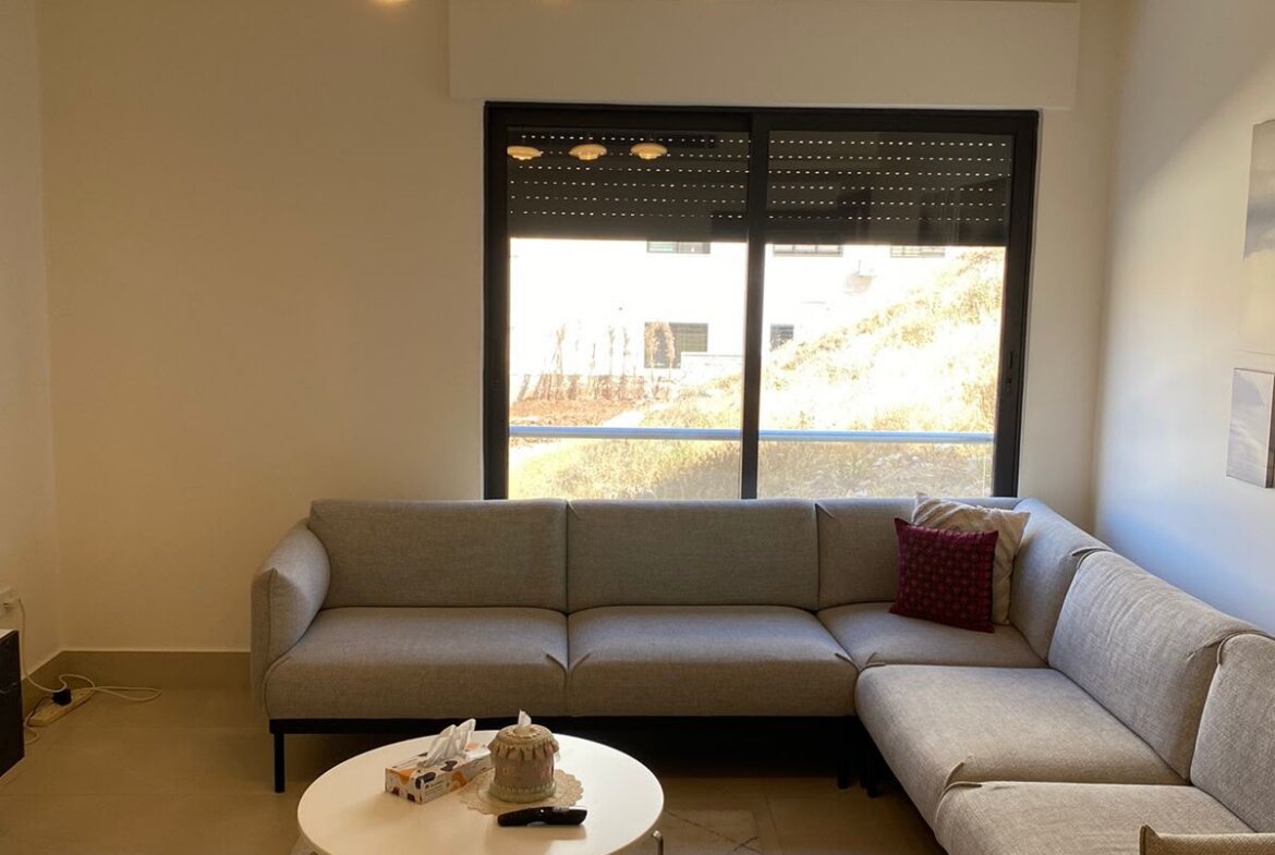 L-shaped beige living room sofa with a red cushion, two round white coffee tables, and a beige rug facing a large window with blinds.