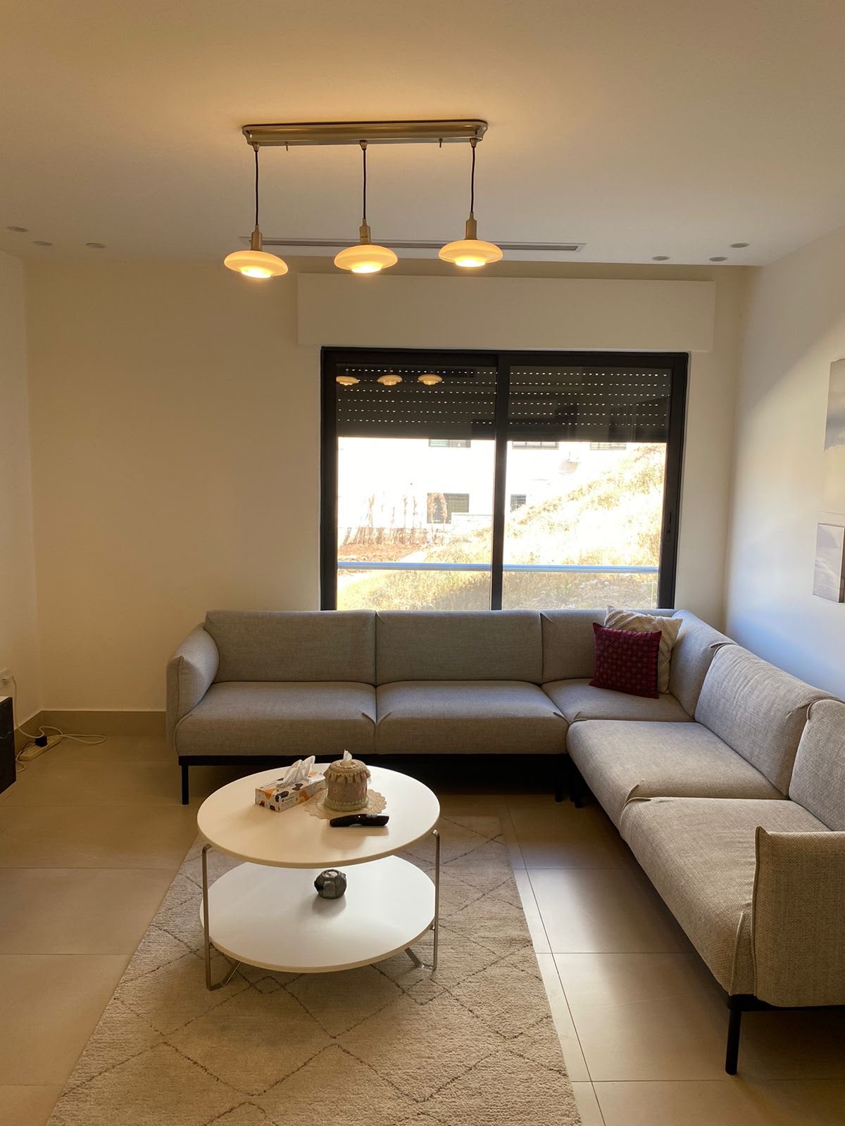 54f68567-fa9a-43d7-86f6-527279724559 L-shaped beige living room sofa with a red cushion, two round white coffee tables, and a beige rug facing a large window with blinds.