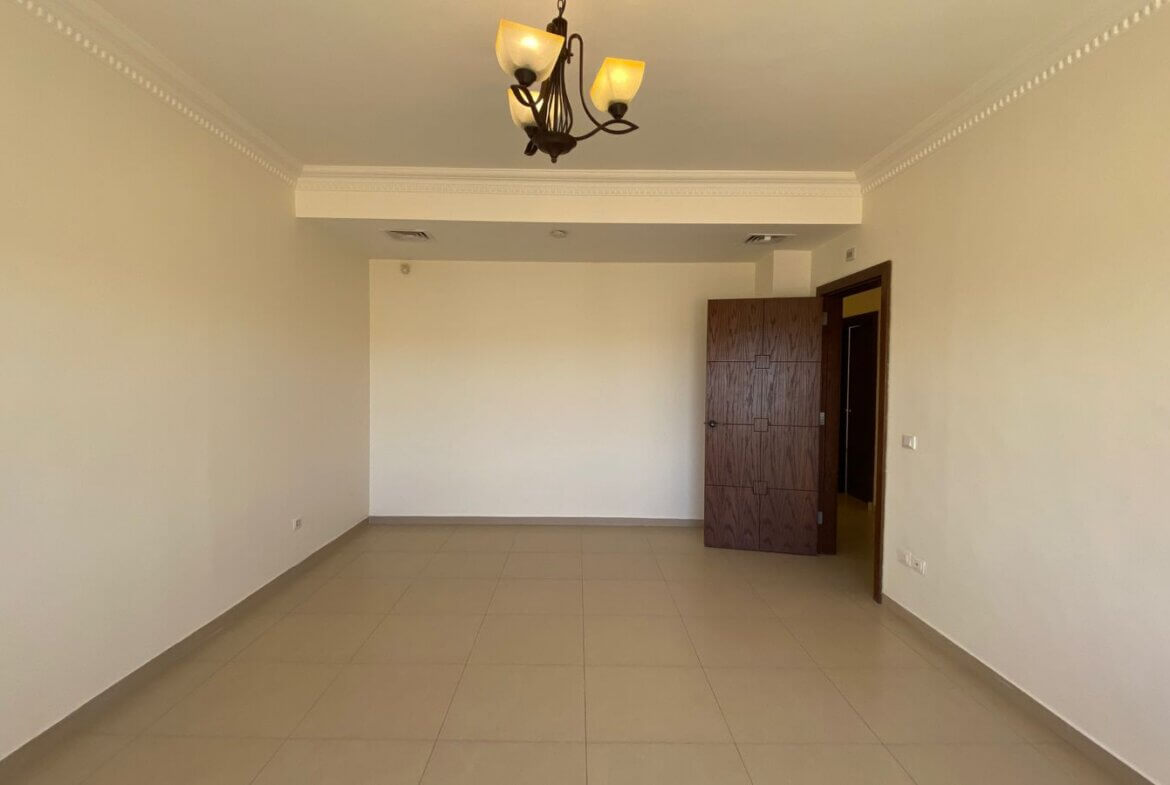 5579f37d-88ba-4886-92f0-04a2d9a5f8f6 Empty, unfurnished room with beige tile floor, white walls, and decorative crown molding; a dark wood chandelier hangs from the ceiling. An open wooden door and a tall wooden cabinet-like panel are visible in the back right corner.