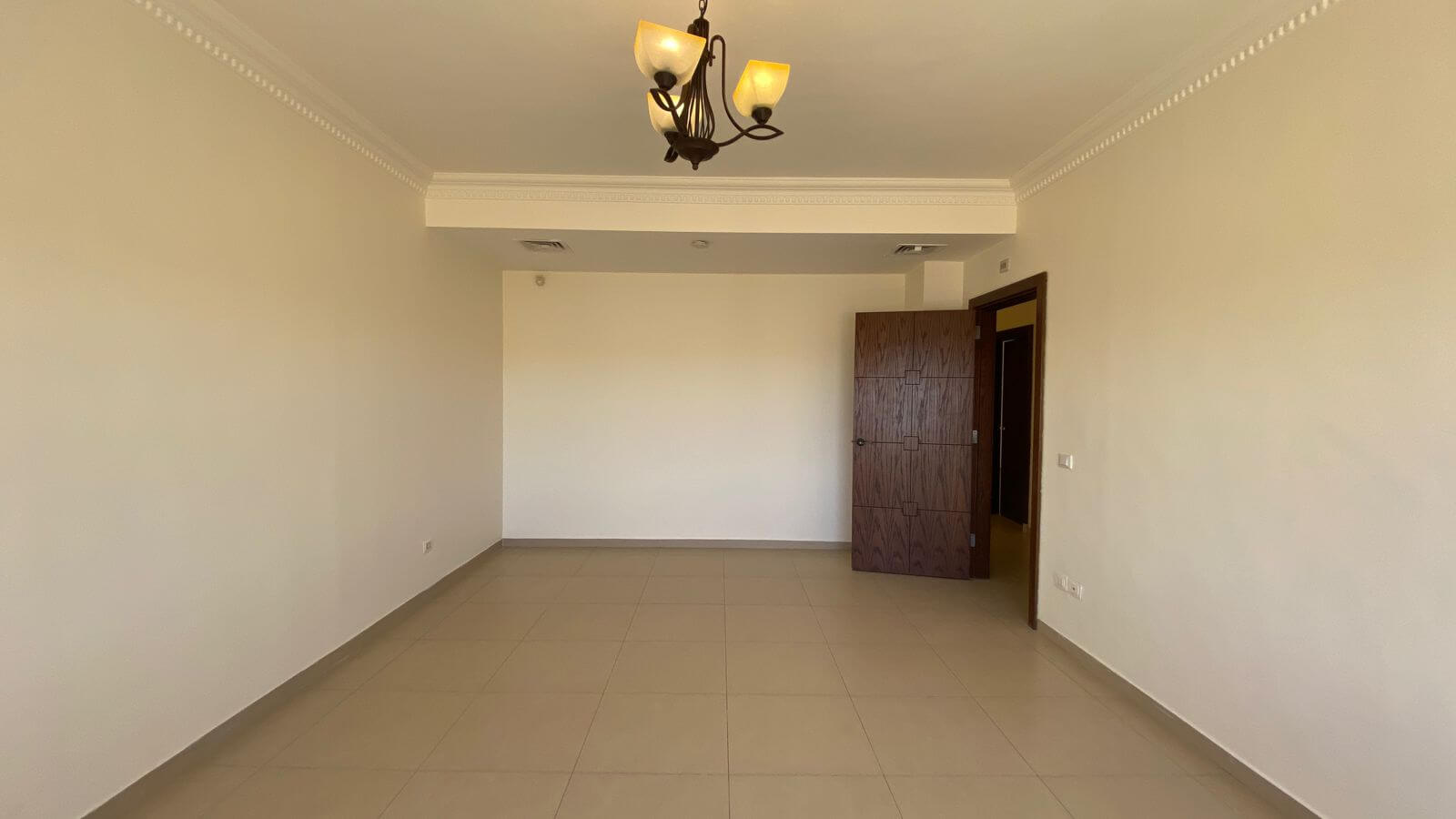 5579f37d-88ba-4886-92f0-04a2d9a5f8f6 Empty, unfurnished room with beige tile floor, white walls, and decorative crown molding; a dark wood chandelier hangs from the ceiling. An open wooden door and a tall wooden cabinet-like panel are visible in the back right corner.
