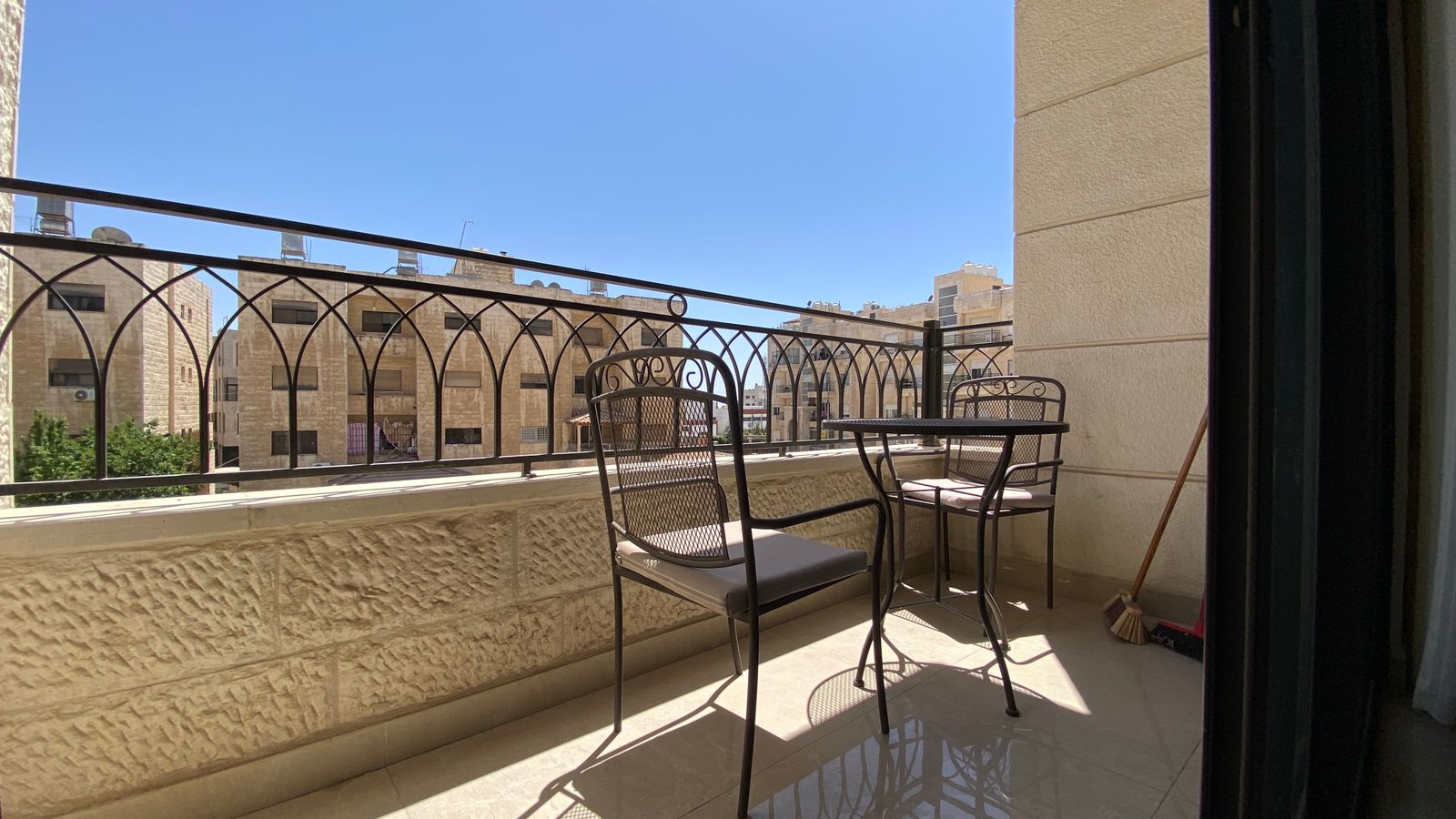 559d04d7-2400-4345-bed1-e21904dbf2d3 Sunlit balcony with a metal patio table and two chairs, overlooking beige stone buildings against a clear blue sky.